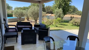 Une terrasse couverte, aménagée avec des meubles en osier, une table en verre et offrant une vue sur un jardin paysager et des arbres au loin.