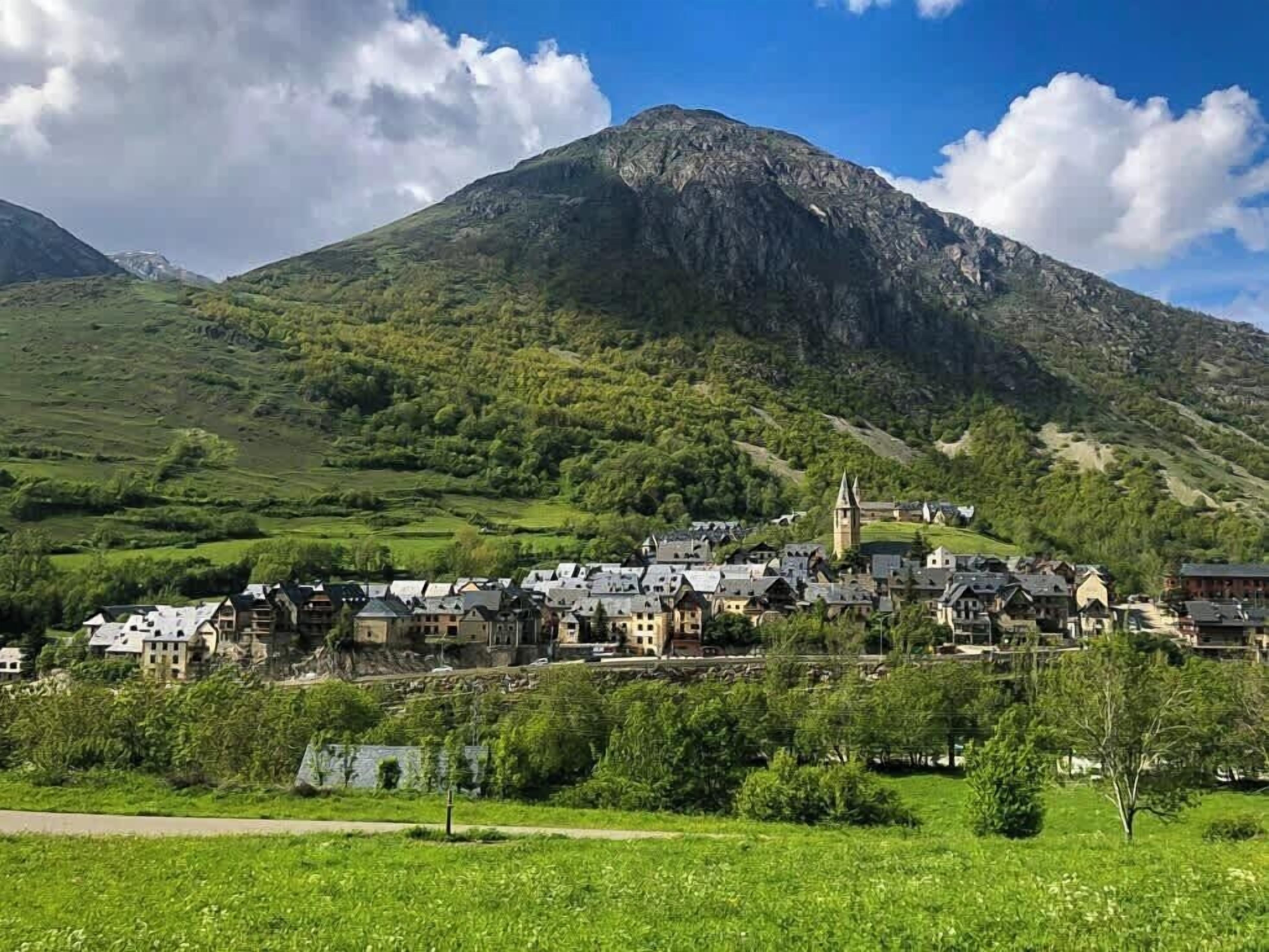Un pueblo con edificios tradicionales ubicado en un valle, rodeado de colinas verdes y una montaña destacada al fondo.