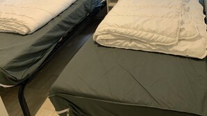Une chambre avec deux lits simples, chacun doté d’un oreiller blanc et d’un matelas de couleur foncée. Une petite table de chevet se trouve entre les lits, et un rideau à motifs est visible à l’arrière-plan.
