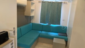 Un espace de vie compact comprenant un canapé d’angle turquoise, une petite table d’appoint blanche, un système de climatisation intégré et un rideau à motifs.