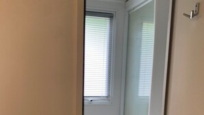 Un couloir étroit mène à une pièce par une porte. La pièce est dotée d’une fenêtre avec des stores, d’un radiateur et d’une porte avec une poignée.