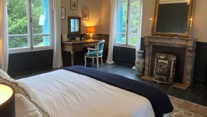 A bedroom with a large bed, a desk, a chair, a chandelier, a fireplace, and large windows.