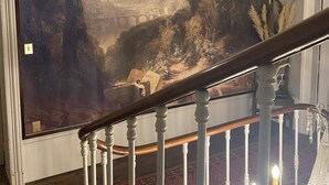 A large landscape painting on a wall, a wooden staircase with white balusters, and a chandelier with candles.