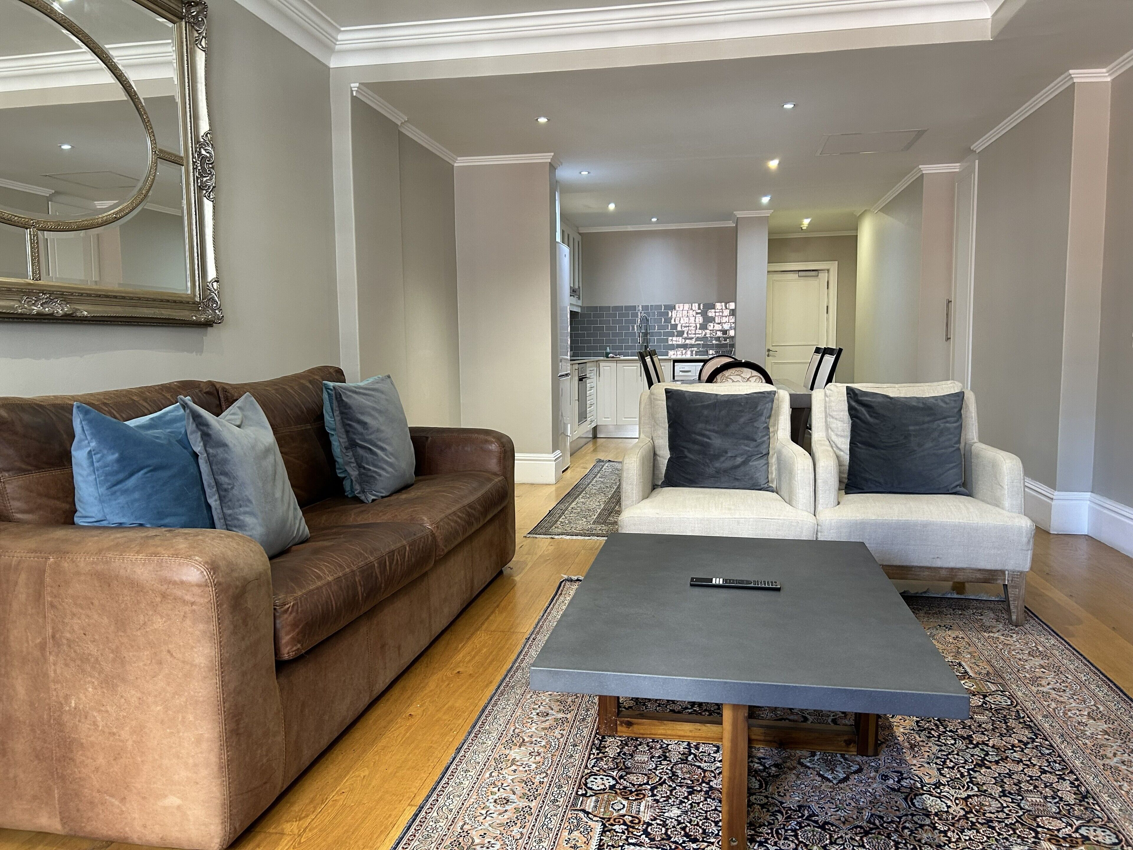 A living room with a brown leather sofa, two white armchairs, a grey coffee table, and a patterned rug.