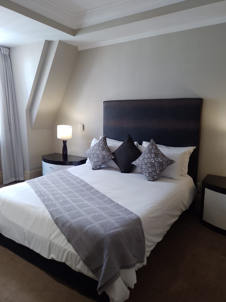A hotel room with a large bed, a nightstand with a lamp, and a view of the sky through a window.