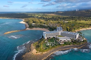 A coastal hotel with multiple buildings, a beach, and wind turbines in the background.
