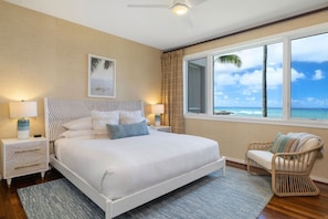 A bedroom with a large bed, two bedside tables, a chair, a window with a view of the ocean, and a ceiling fan.