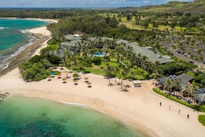 An aerial view of a resort with a beachfront, palm trees, and a golf course in the background.