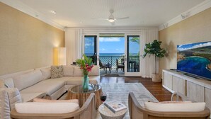 A spacious living room with a large sofa, a coffee table, and a TV on a wooden cabinet. There is a balcony with a view of the ocean. - Presidential Suites • Turtle Bay 4BR Oceanfront Villa (Kahuku)