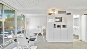 A modern, minimalist room with a glass sliding door leading to an outdoor area, a glass dining table with chairs, a bed with white bedding, and a white cabinet with kitchen appliances. - 1957 Modern Hideaway - A Ryson Property (Palm Springs)