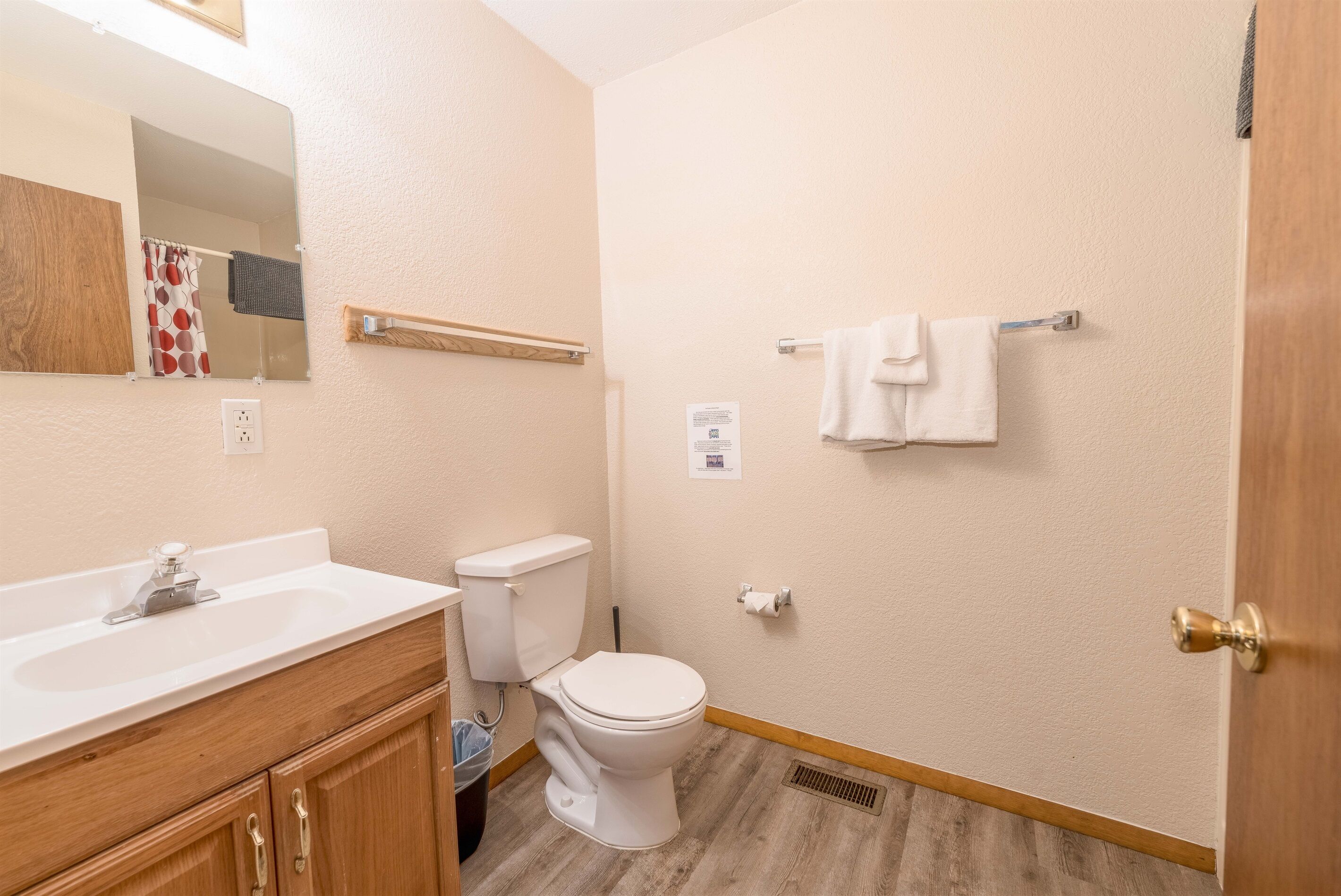 Un bagno piccolo con lavandino, wc e specchio bianchi. C'è un portasciugamani sulla parete e un pavimento in legno.
