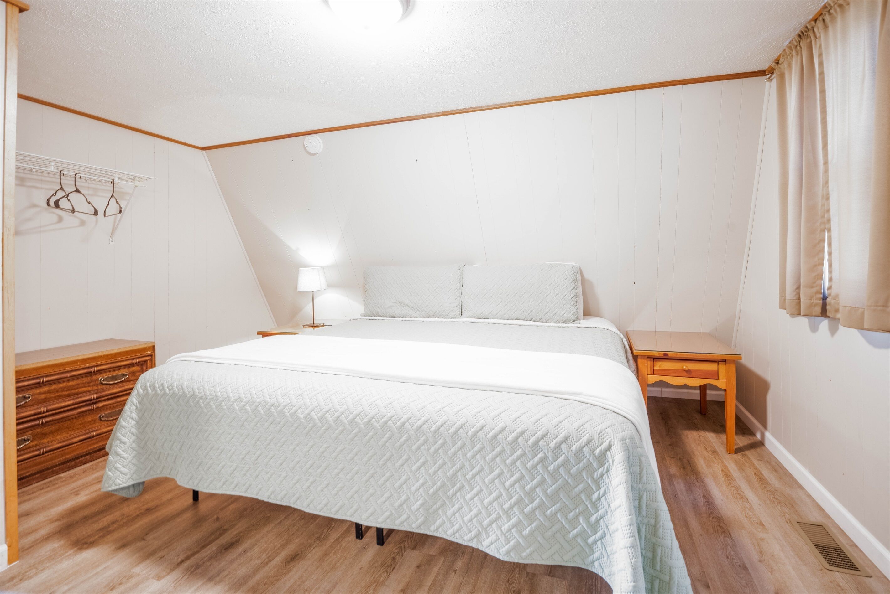 Een slaapkamer met een bed, houten nachtkastjes, een dressoir en een wandlamp.