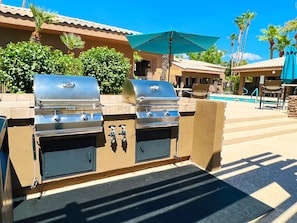 Uma área de churrasco ao ar livre com duas churrasqueiras a gás embutidas, um espaço de estar junto à piscina e um guarda-sol coberto.