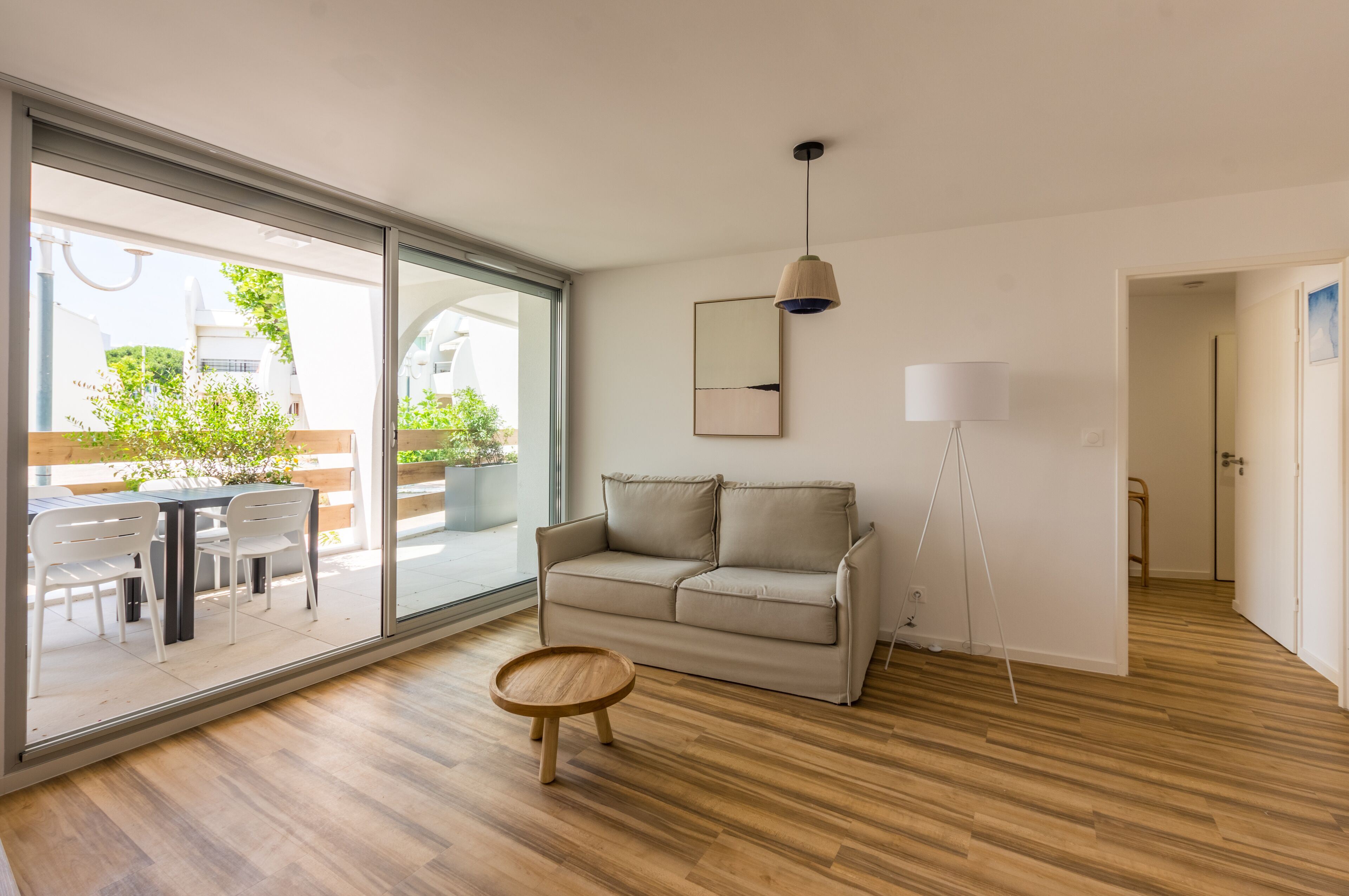 Una sala de estar moderna con un ventanal corredizo que da a un espacio exterior, un sofá color beige, una pequeña mesa de madera y una lámpara colgante.