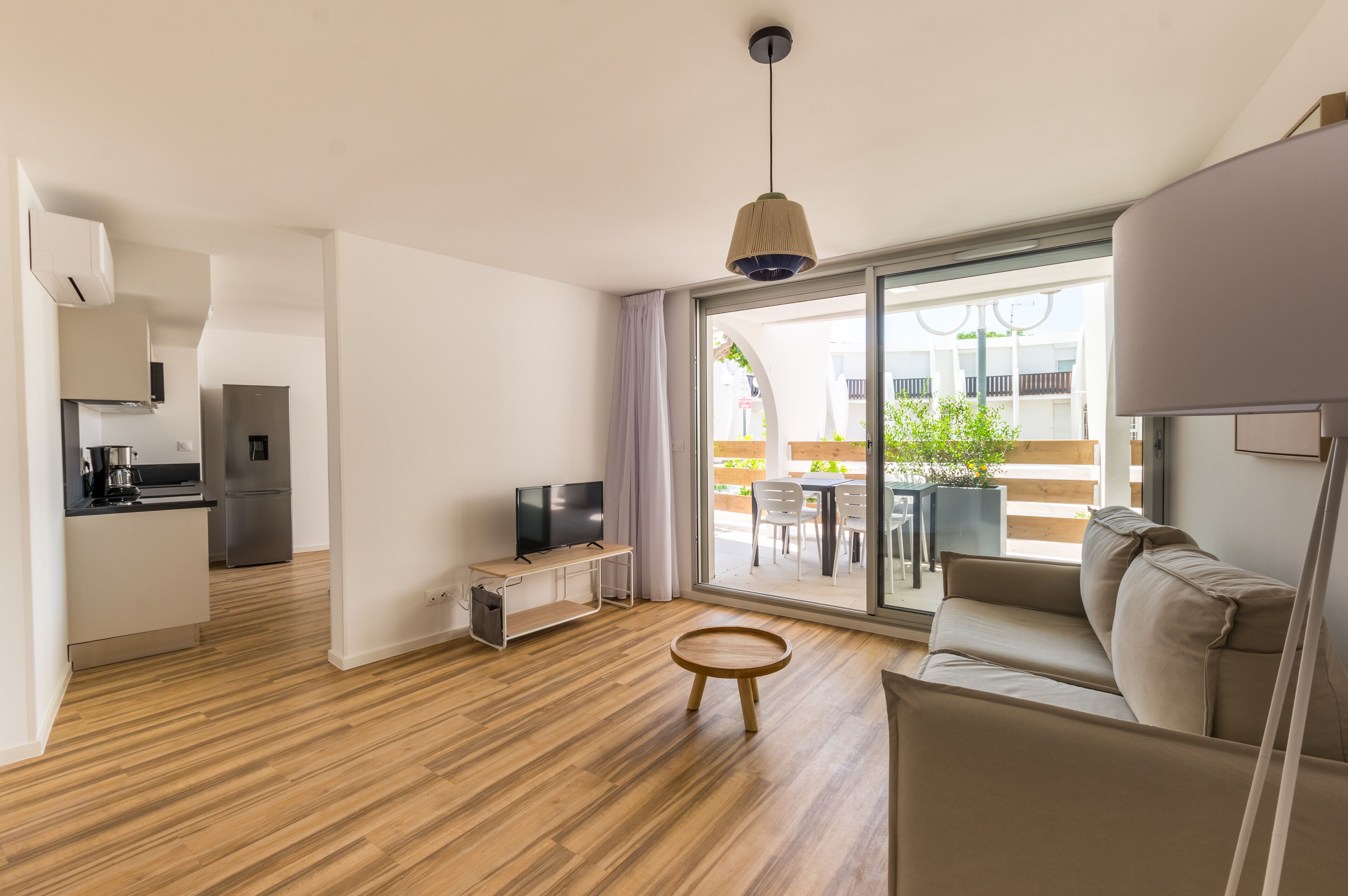 Una sala de estar moderna con un sofá, un televisor y piso de madera. Hay un ventanal corredero que da a un espacio exterior con una mesa y sillas.