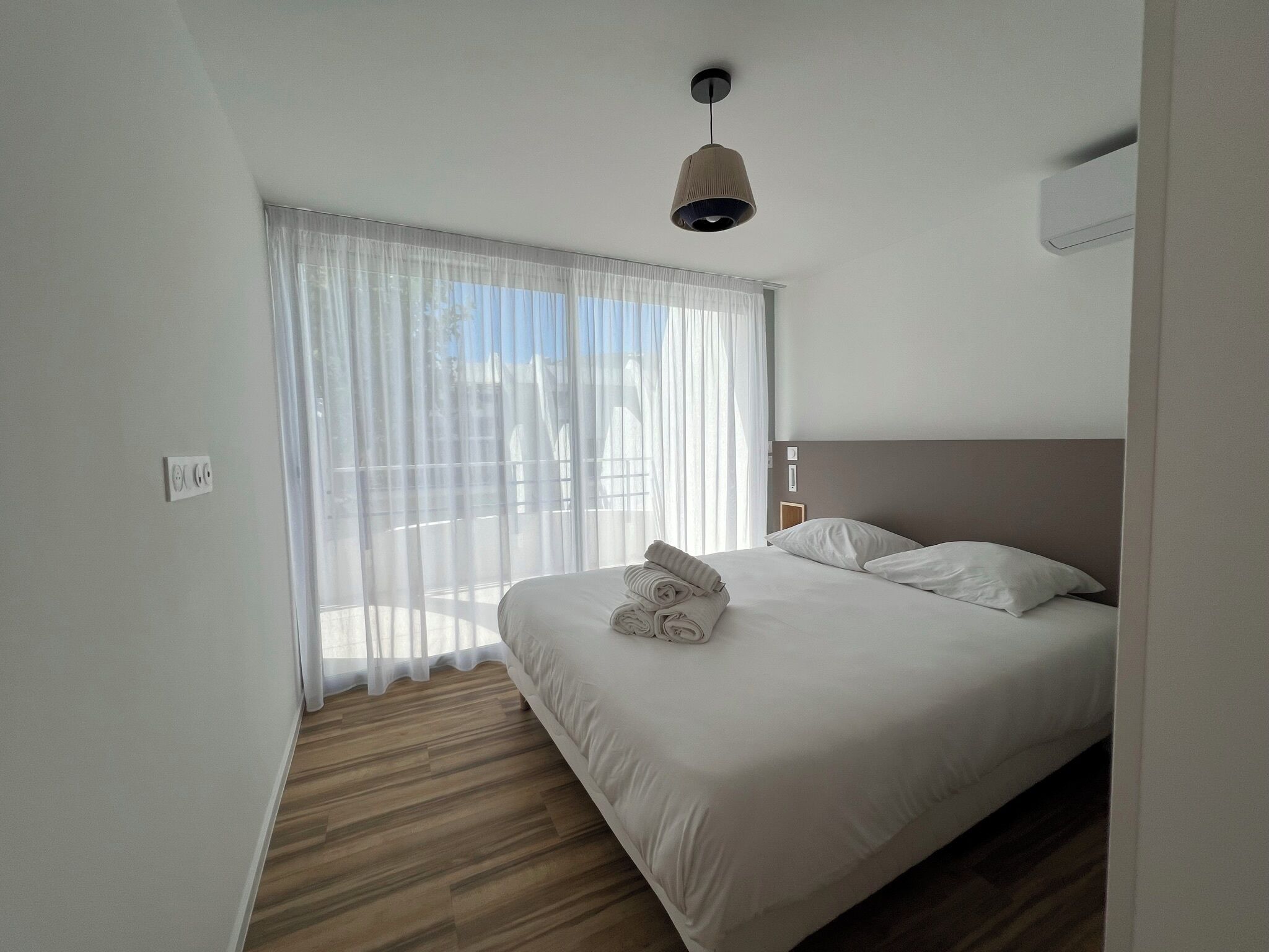 A hotel room with a bed, a window with sheer curtains, a wooden floor, and a ceiling light.