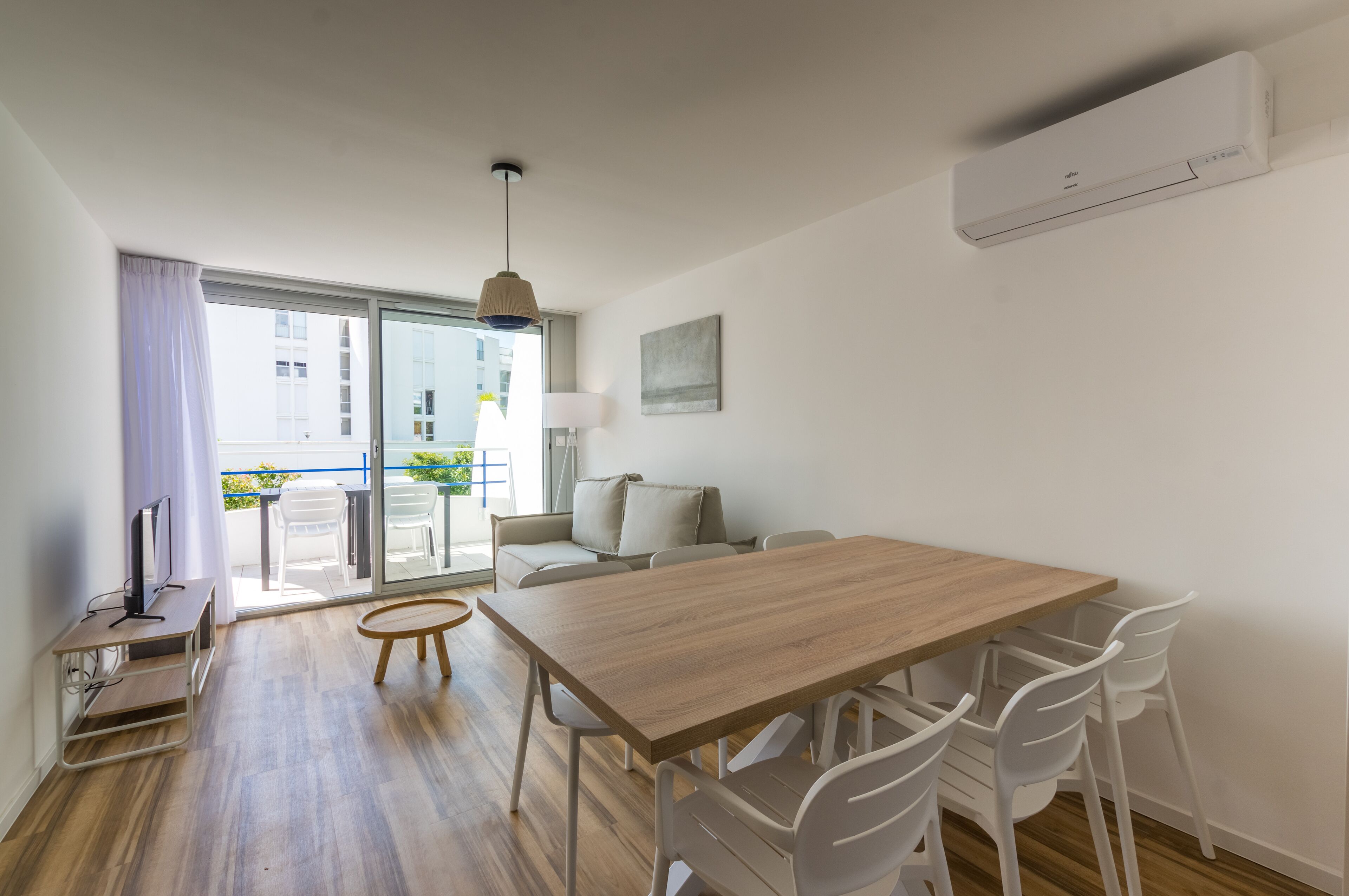 Una sala de estar moderna con una mesa de comedor de madera, sillas blancas, un sofá, un televisor y un amplio ventanal con vistas a edificios.