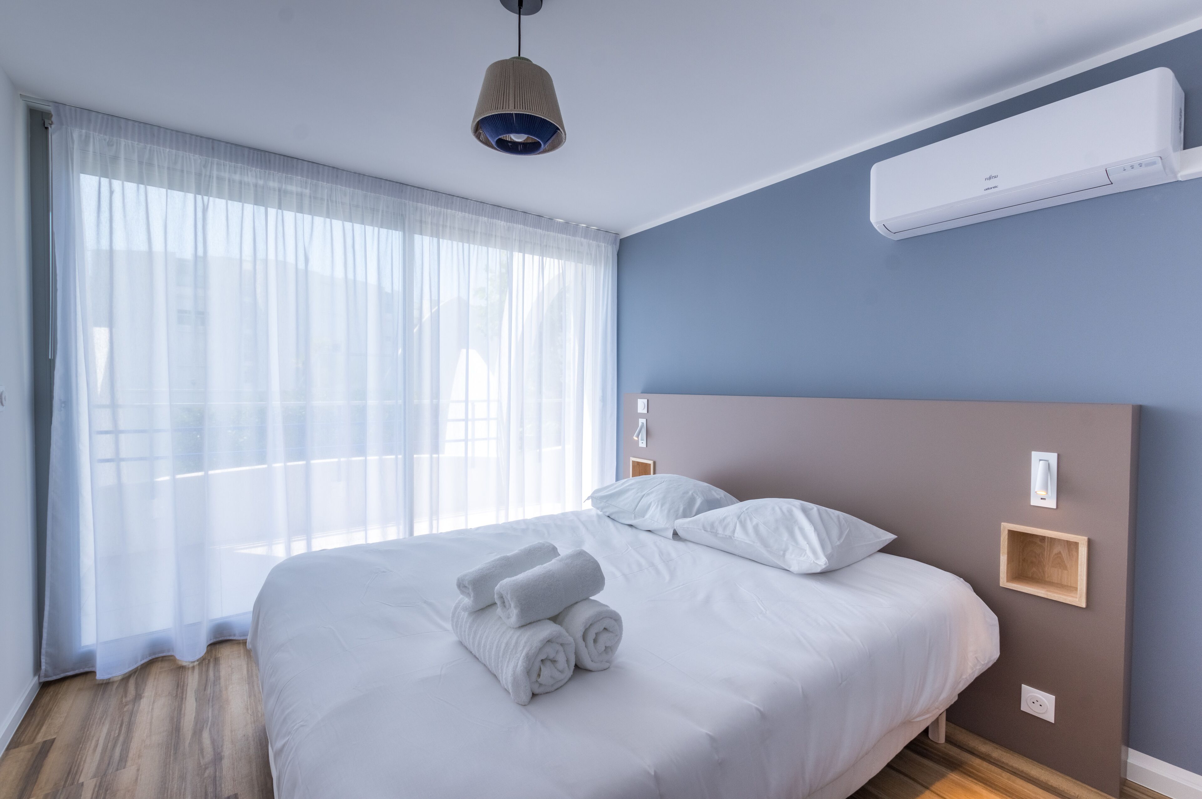 A neatly made bed with rolled white towels, a ceiling-mounted air conditioner, and a bedside table with a light fixture.