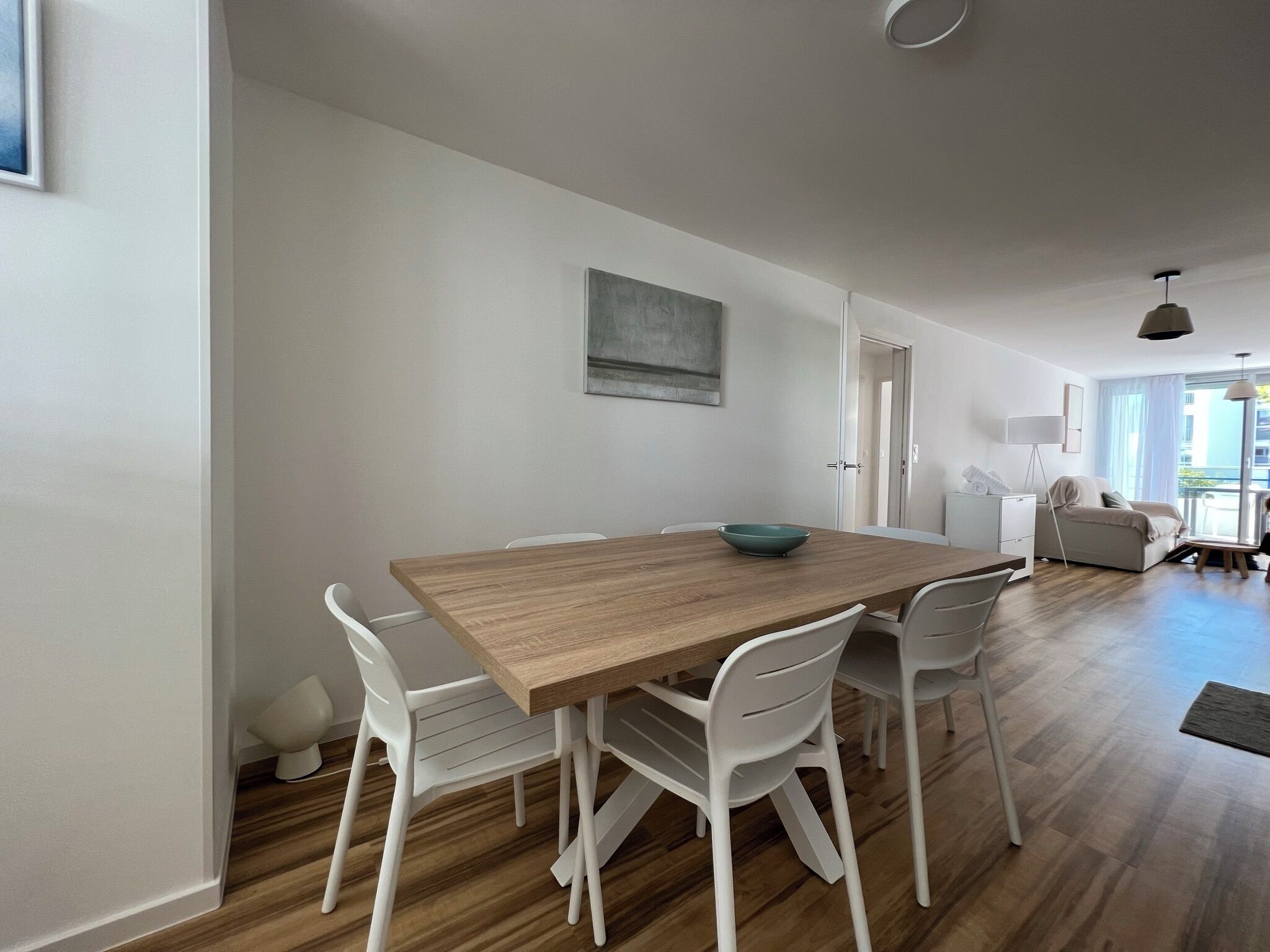 A modern dining area with a wooden table and white chairs, a bowl on the table, and a painting on the wall.