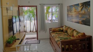 A living room with a sofa, a TV, a ceiling fan, and a view of palm trees outside. - Apartamento Térreo, no Local Mais Estratégico de Ubatuba!!! (Ubatuba)