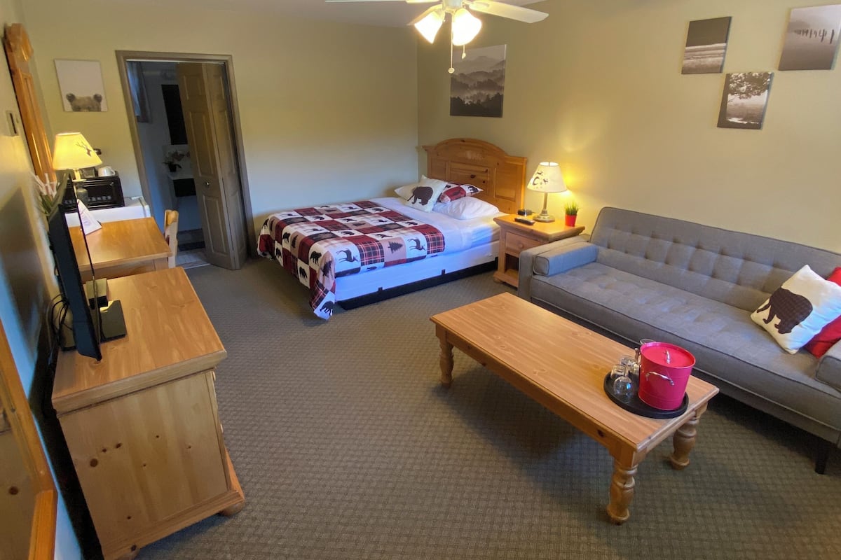 A room with a bed, a sofa, a wooden table, a ceiling fan, and a desk with a lamp.