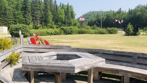 Een houten terras met een vuurkuil, omringd door een gazon en bomen.
