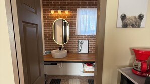 Een badkamer met betegelde vloer, een wastafel, een spiegel, een raam met gordijn en een rode koffiezetapparaat op het aanrecht.