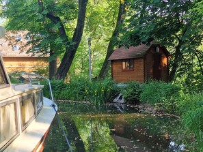 A wooden cabin with a small window and a door, surrounded by lush greenery and a pond.