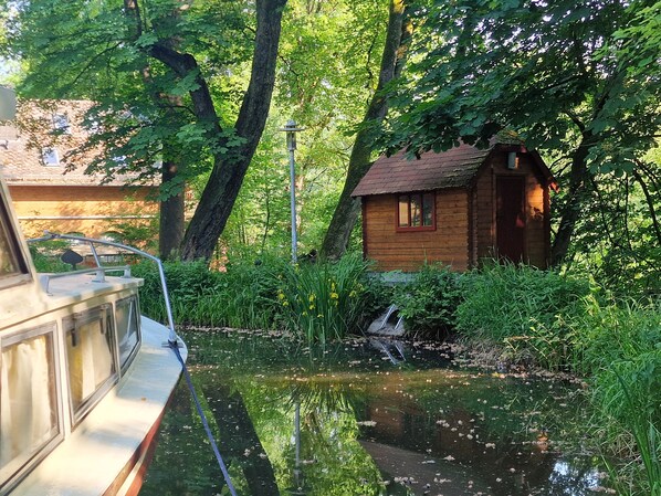 Eine Holzhütte mit kleinem Fenster und Tür, umgeben von üppigem Grün und einem Teich.