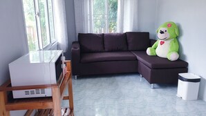 Una sala de estar con un sofá marrón oscuro, un peluche verde, un electrodoméstico blanco y un cubo de basura.