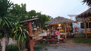 Un área de comedor al aire libre con mesas y bancos de madera, rodeada de exuberante follaje y una estructura de techo de paja.