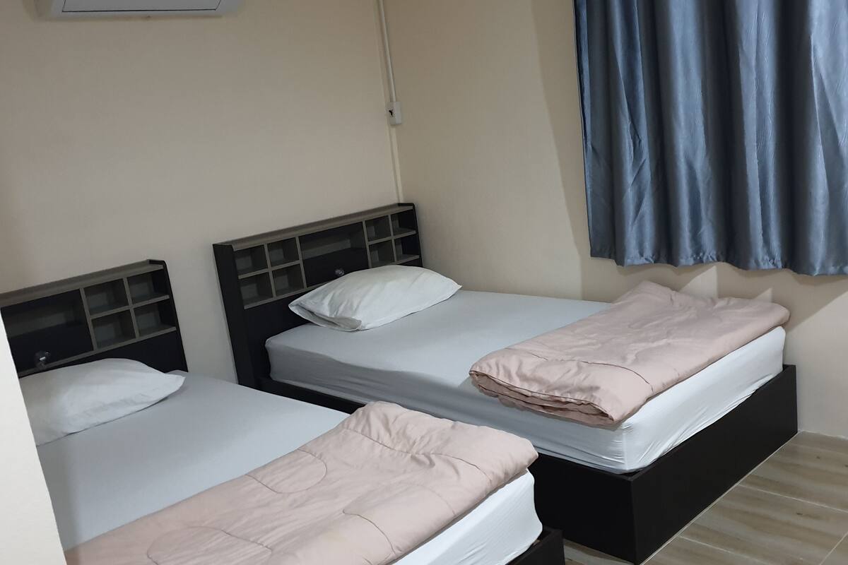 Two single beds with headboards, a wall-mounted air conditioner, and a window with blue curtains.