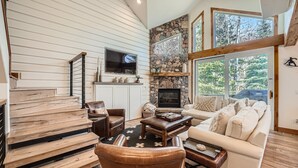 A cozy living room with a stone fireplace, wooden staircase, and large windows offering a view of the outdoors. - 1890 Lions Ridge Loop, #9-West Vail Remodeled Home, Hot Tub, On Bus Route, Close to Vail! (Vail)
