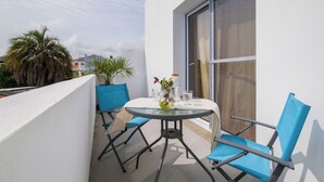 A balcony with a round glass table, two blue chairs, a potted plant, and a window with a curtain. - Botanic Garden Duplex (Ponta Delgada)