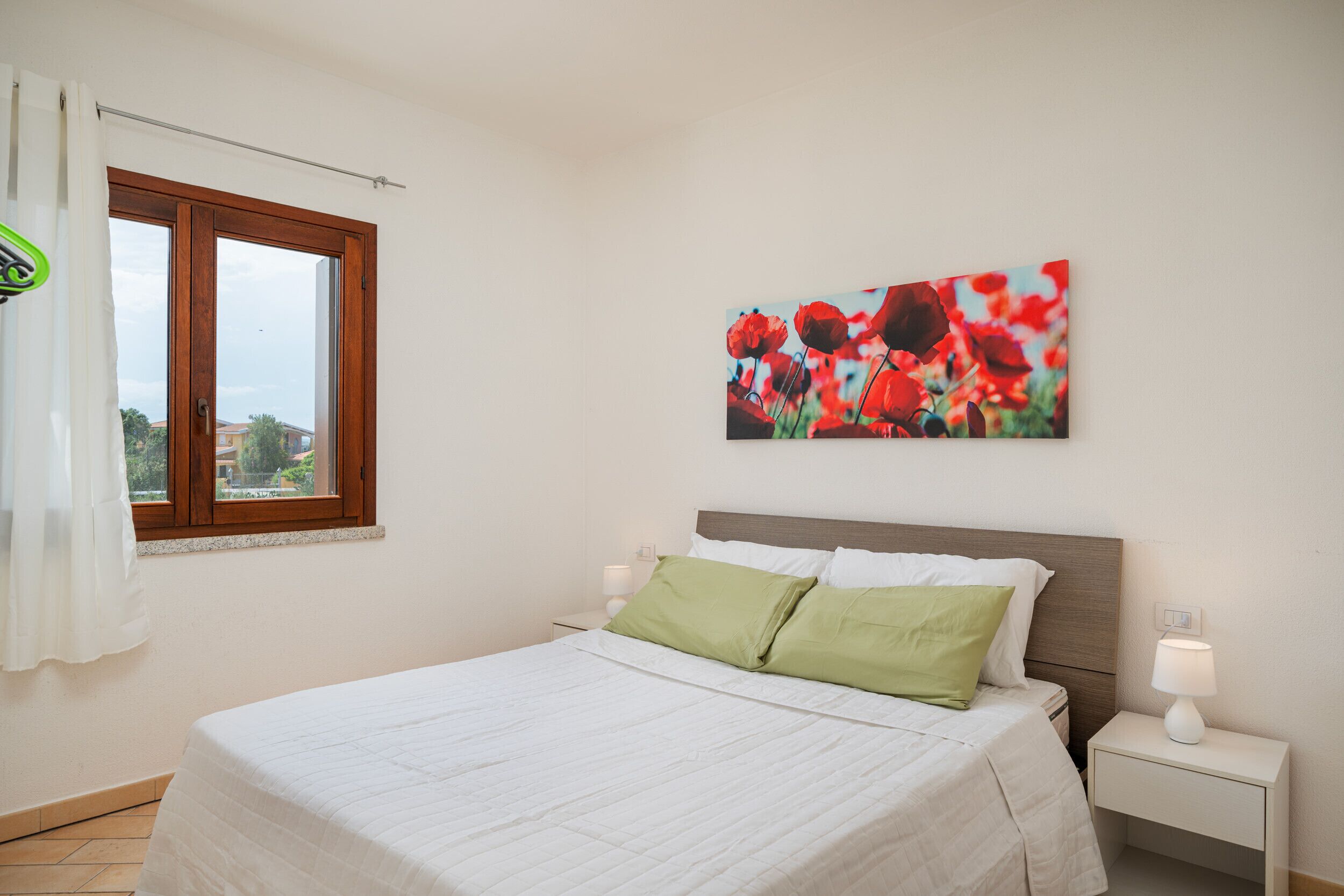 A bedroom with a bed, a nightstand, a lamp, a window with curtains, and a painting on the wall.