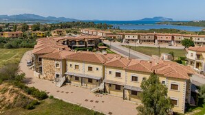 A coastal residential area with terracotta-roofed buildings, a paved road, and a view of the sea and mountains in the distance.
