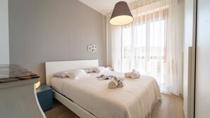 Un letto rifatto con cura, con testiera bianca, una lampada a sospensione e vista su un balcone attraverso tende leggere.