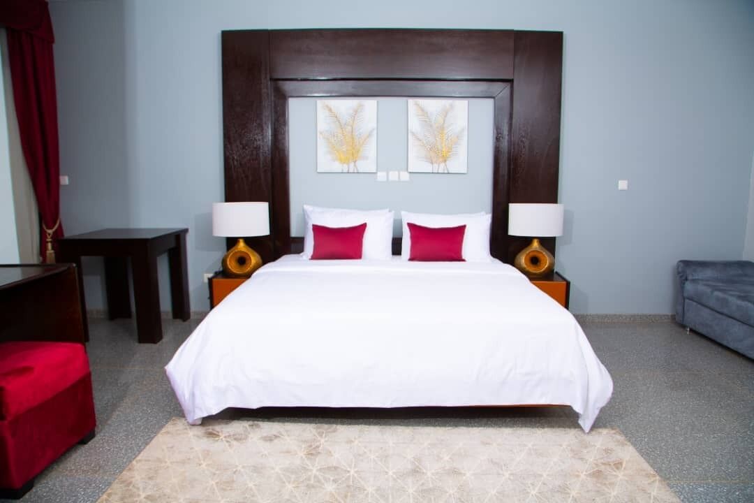 A neatly arranged bedroom with a large bed, two bedside tables, a red ottoman, and a grey sofa.