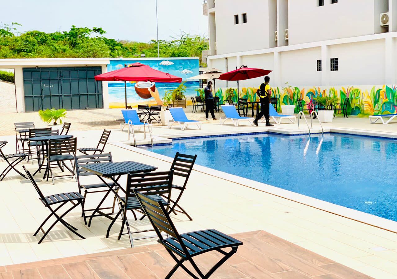 Une piscine entourée de chaises longues et de tables, avec une fresque murale et un bâtiment doté d’un toit plat.