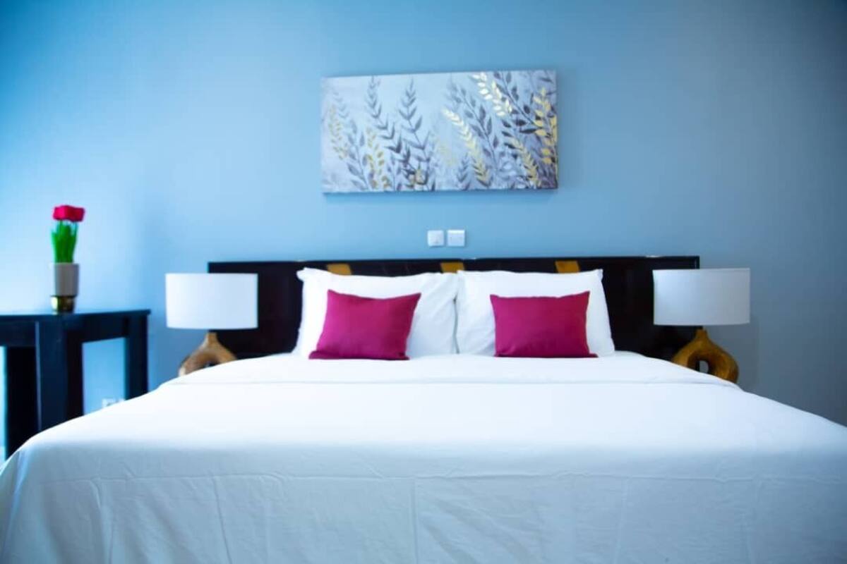 A hotel room with a bed, two bedside lamps, a painting on the wall, and a vase with a rose on a nightstand.