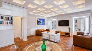 Uma sala de estar espaçosa com piso de madeira em padrão herringbone, paredes brancas e teto com iluminação embutida. Há uma televisão de tela plana grande fixada na parede, um sofá de couro marrom e uma mesa de centro branca com um vaso verde.
