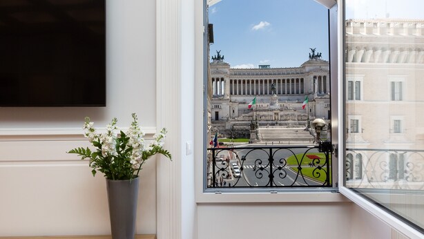 Quarto com uma grande janela com vista para um edifício histórico, uma planta em vaso e uma televisão de tela plana na parede.