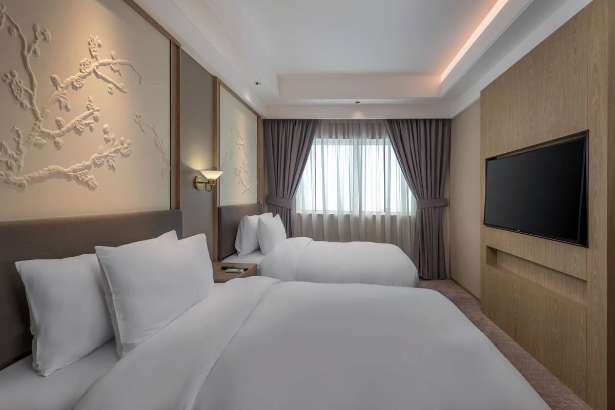 Ein Hotelzimmer mit zwei Betten, einem Flachbildfernseher, einer Wandlampe und einem großen Fenster mit Vorhängen.