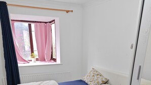 Une chambre avec un lit simple, une tête de lit blanche, une couvre-lit bleue et un coussin à motifs. On aperçoit une fenêtre avec des rideaux roses et une plante en pot.