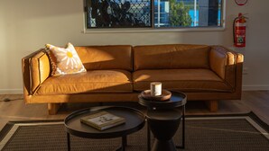 A brown leather sofa with a cushion, a small round table with a book, and a window with a view of buildings and a fire extinguisher on the wall. - Mackay Adventure Lodge (Andergrove)