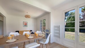 Un coin repas avec une table en bois dressée pour quatre personnes, des chaises blanches et une vue sur l’extérieur, accessible par une porte vitrée.