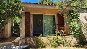Una casa rústica con una puerta corredera de vidrio, un pequeño patio con mesa y sillas, y un jardín con diversas plantas y árboles.