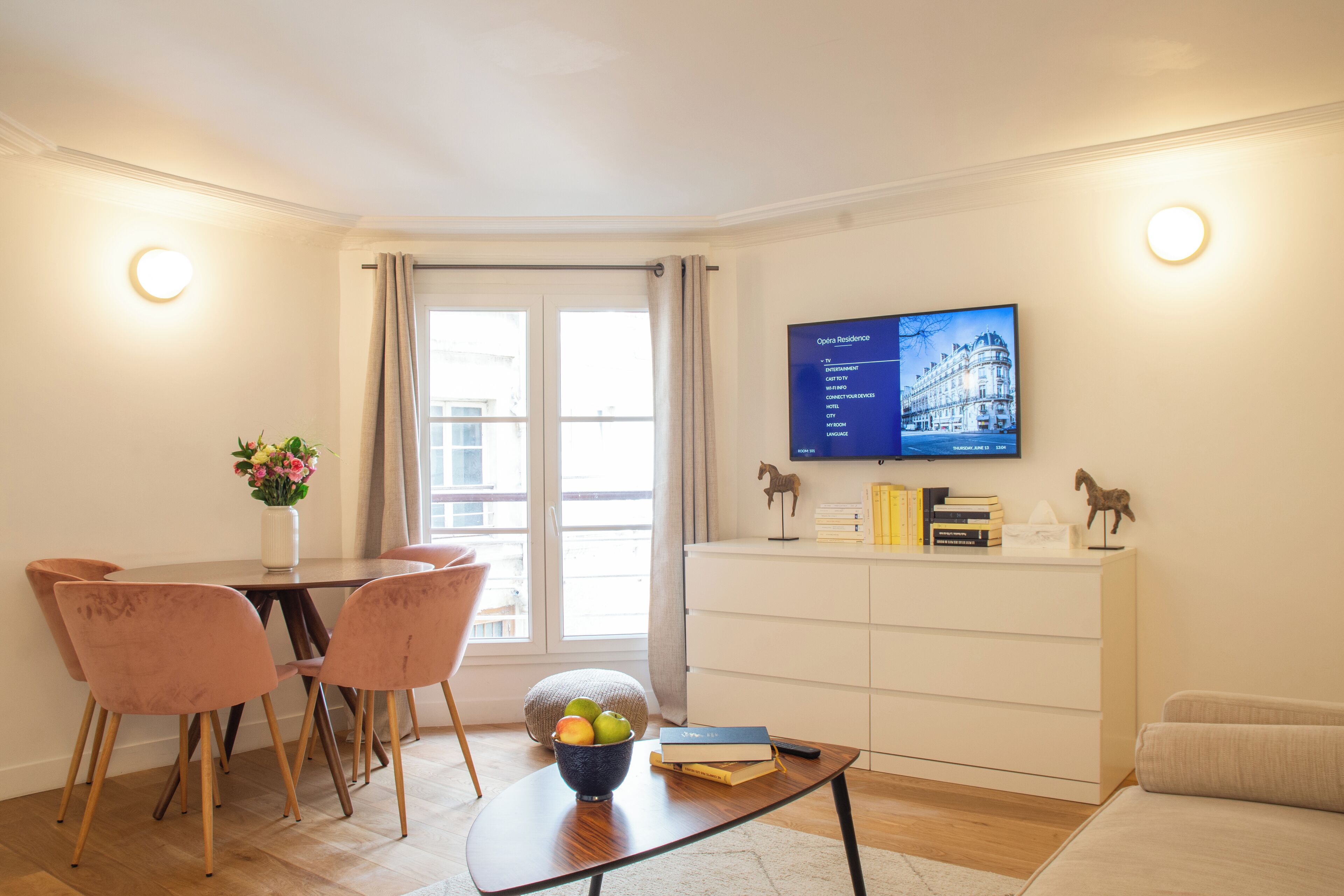 A modern living room with a dining area, a flat-screen TV mounted on the wall, a white cabinet, and a window with curtains.