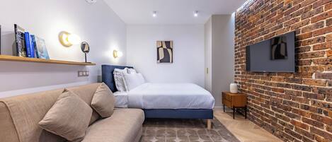 A modern bedroom with a brick wall, a bed with a blue headboard, a beige sofa, a small side table, and a mounted TV.