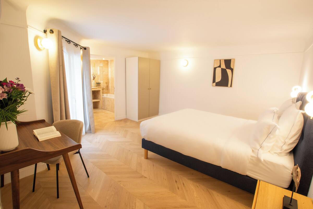 A hotel room with a bed, a desk, a chair, a potted plant, and a bathroom visible through an open door.